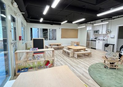Childcare learning space with wooden tables, play corner, and kitchen area for children to explore and learn.