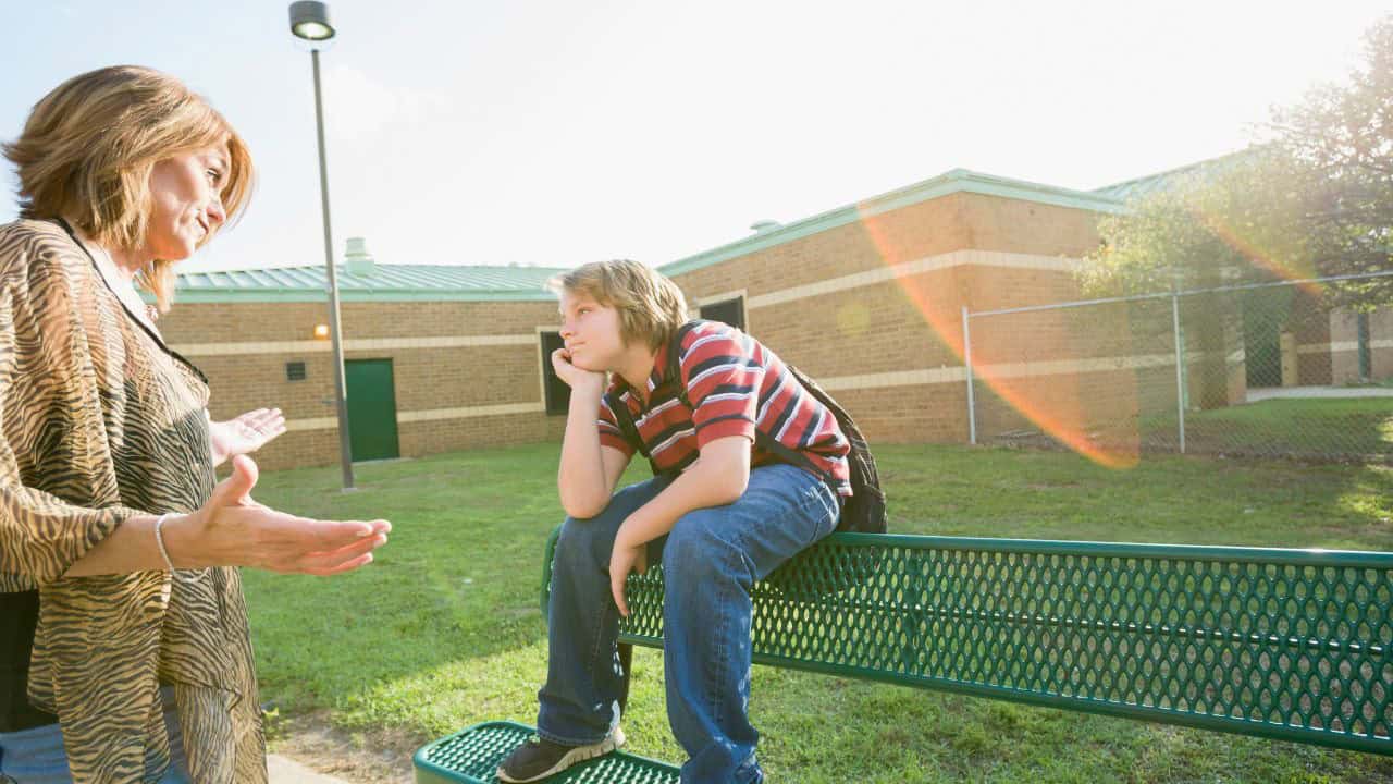 Learning Center Kids Scene. Young boy talking with caregiver outdoors at childcare center, sunny day, school building in background.